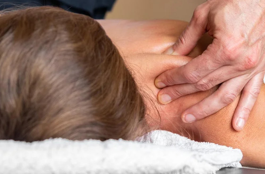 Imagem de um terapeuta a aplicar massagem terapêutica nas costas de uma pessoa deitada numa maca. A prática é focada em aliviar os sintomas somatizados por experiências e emoções do dia a dia, promovendo o equilíbrio físico e energético. O ambiente tranquilo, com plantas ao fundo, reforça a ideia de bem-estar e harmonia.