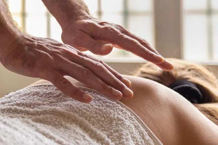 Imagem de um terapeuta a praticar Reiki, com as mãos suspensas sobre as costas de uma pessoa deitada e coberta por uma toalha branca. O Reiki promove a cura energética ao trabalhar no corpo emocional e subtil, ajudando a aliviar manifestações emocionais e a restaurar o equilíbrio interior. O ambiente transmite serenidade, com um fundo decorado por plantas que reforçam a sensação de harmonia.